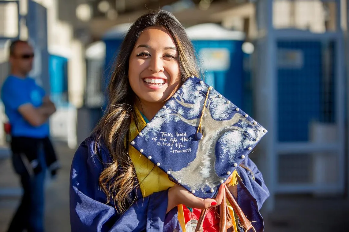 A female student in graduation robes is smiling as she holders her cap up to show a stylized treat painted onto it