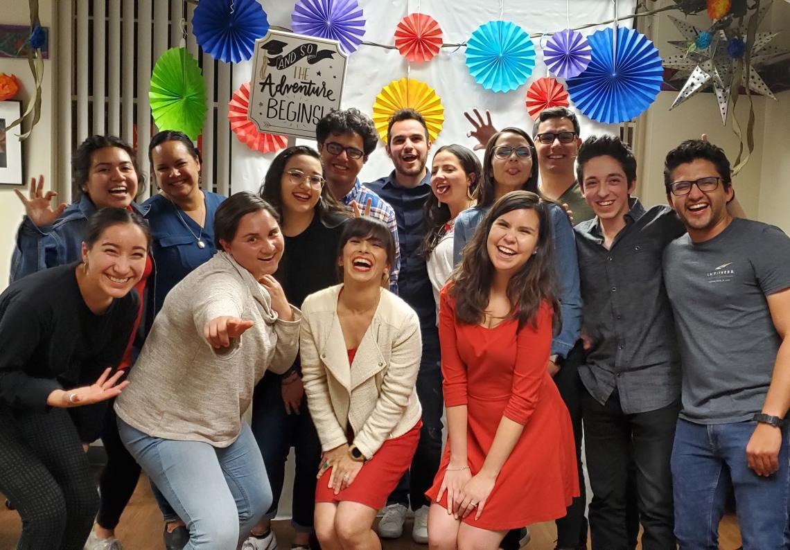 A large group of students smile and laugh as they pose for a photo at the Guerrero Center, traditional hispanic paper decorations can be seen in the background