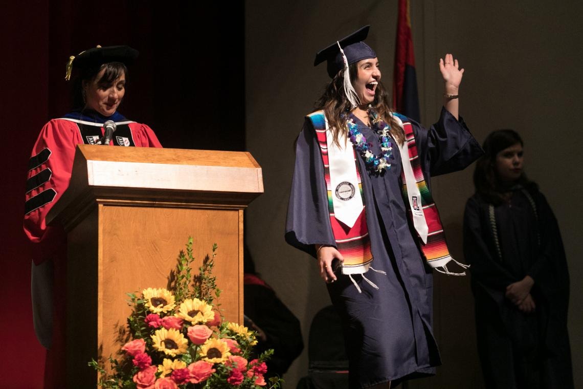 A student waves to the large crowd in Centennial Hall as they gather for the Guerrero Center Convocation, the student is wearing graduation regalia on stage