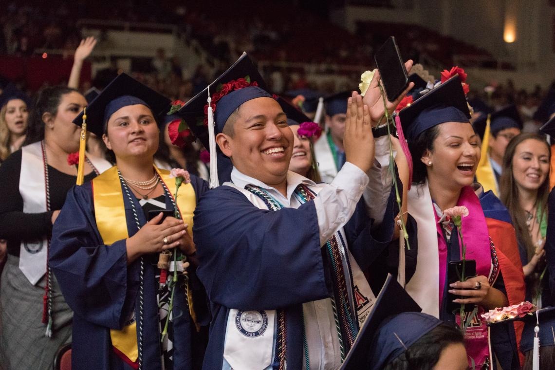 A crowd of students in graduation robes clap their hands at the start of Guerrero Center Convocation held in Centennial Hall, a large crowd is seen in the background
