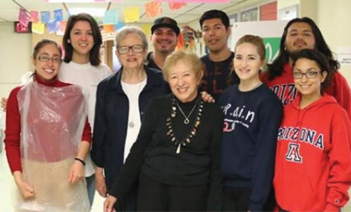 Photo of a group of students and staff as they pose with an abuelita at the start of an AROMAS event, they are all facing the camera and smiling