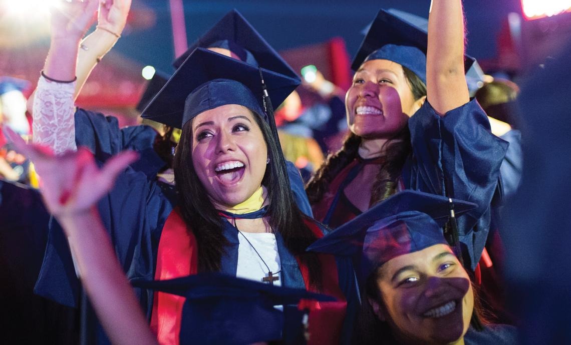 A group of students in graduation regalia celebrate with smiles and their hands in the air at the end of the Commencement Ceremony